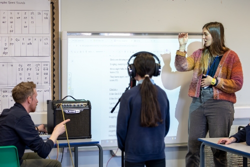 Classroom scene including a primary school pupil wearing headphones near a whiteboard, and two musicians pointing at equipment and the board.