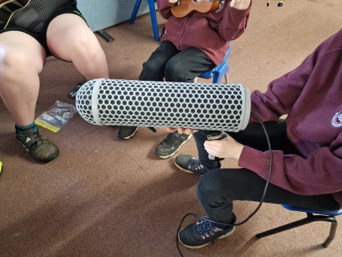 Three people seated indoors, one holding a large microphone and another playing a Ukulele.  Upload