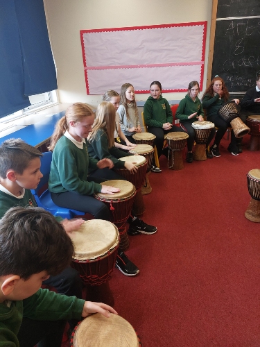 Primary Pupils seated in a semi-circle in a classroom, each playing a drum during a Drumfun Session.