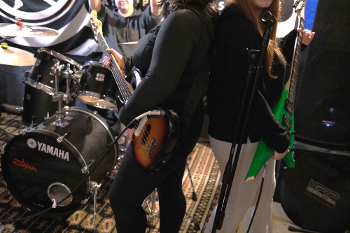 Two young people standing back-to-back with electric guitars in front of a Yamaha drum set, with music equipment visible indoors.