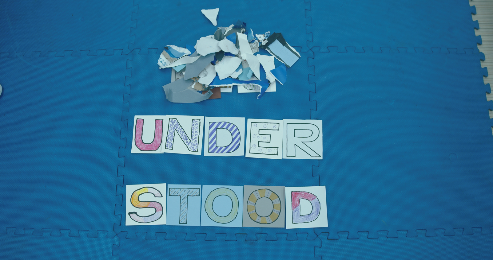 Decorated letters spelling “UNDERSTOOD” on a blue foam mat, with torn paper pieces above.
