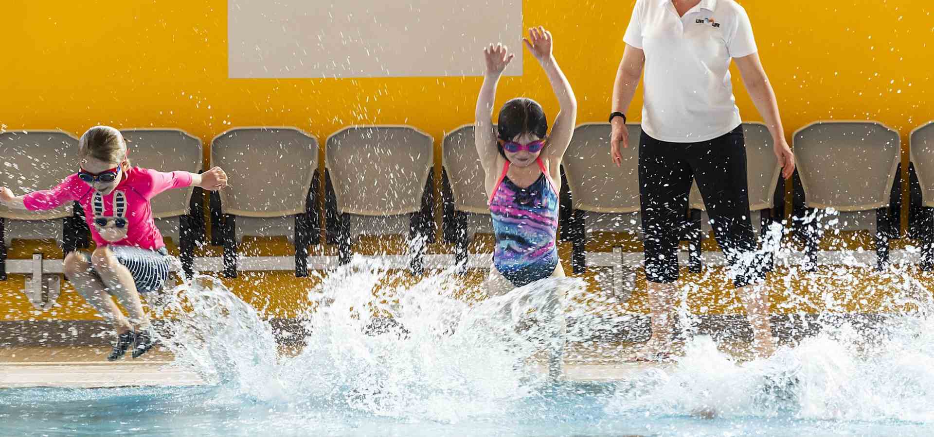 two children jumping into the pool as the teacher watches at the side