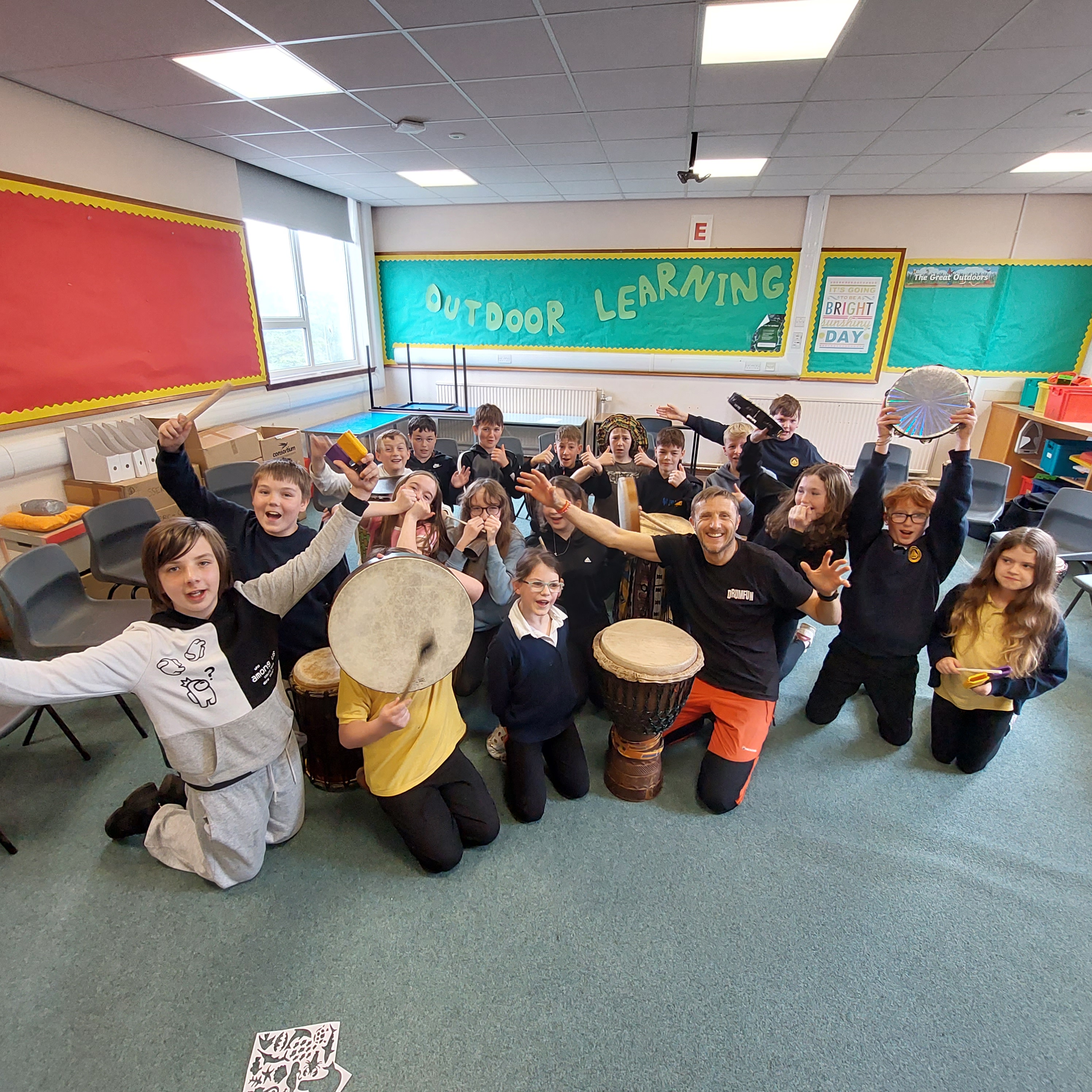 Classroom group photo with pupils and a tutor, some holding drums, and others with their hands up.  They are all smiling.