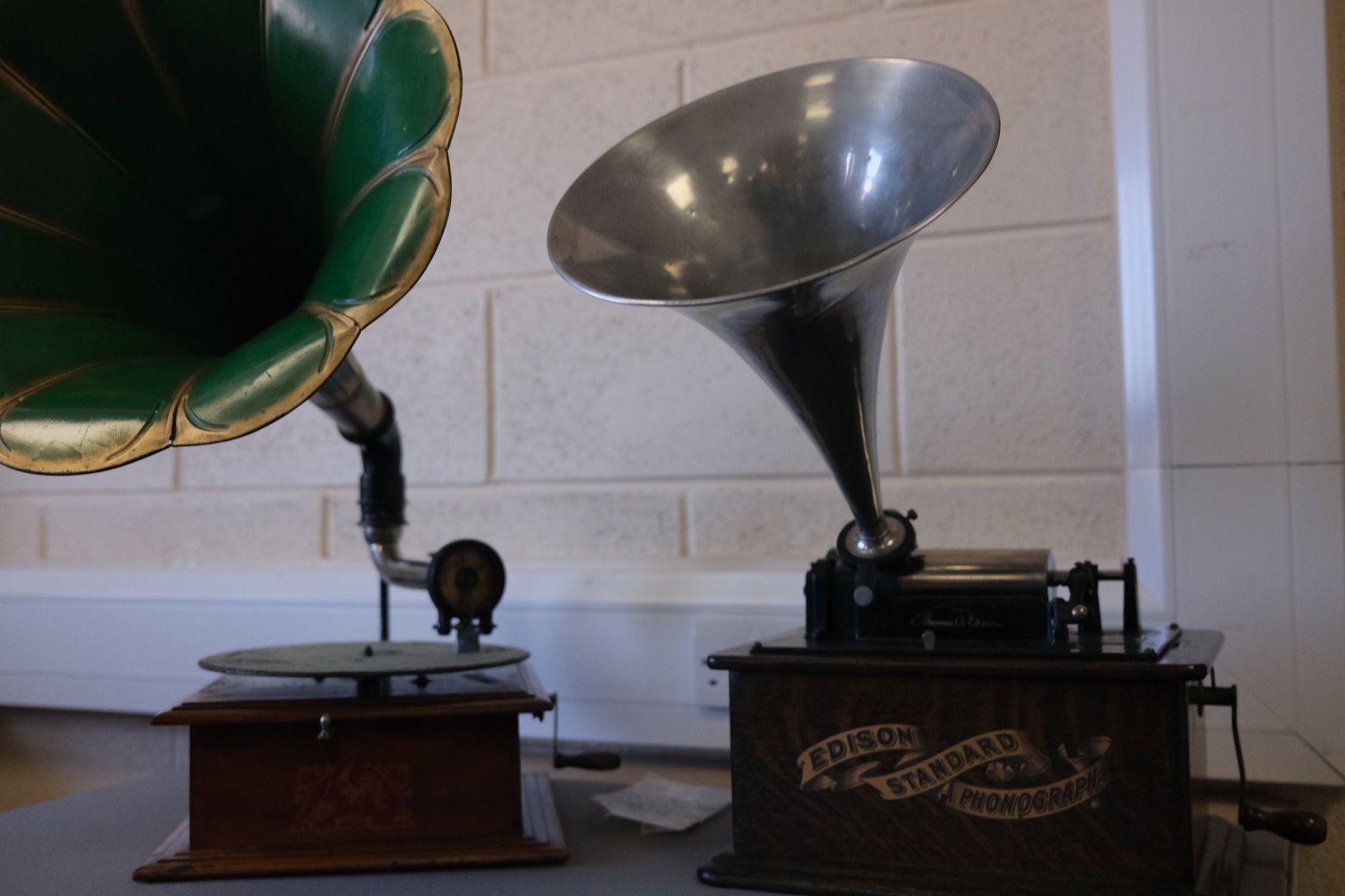 Two antique Edison Standard Phonographs on a table, one with a green horn and the other with a silver horn. The yellow object is a flared horn used to amplify sound from the phonograph.