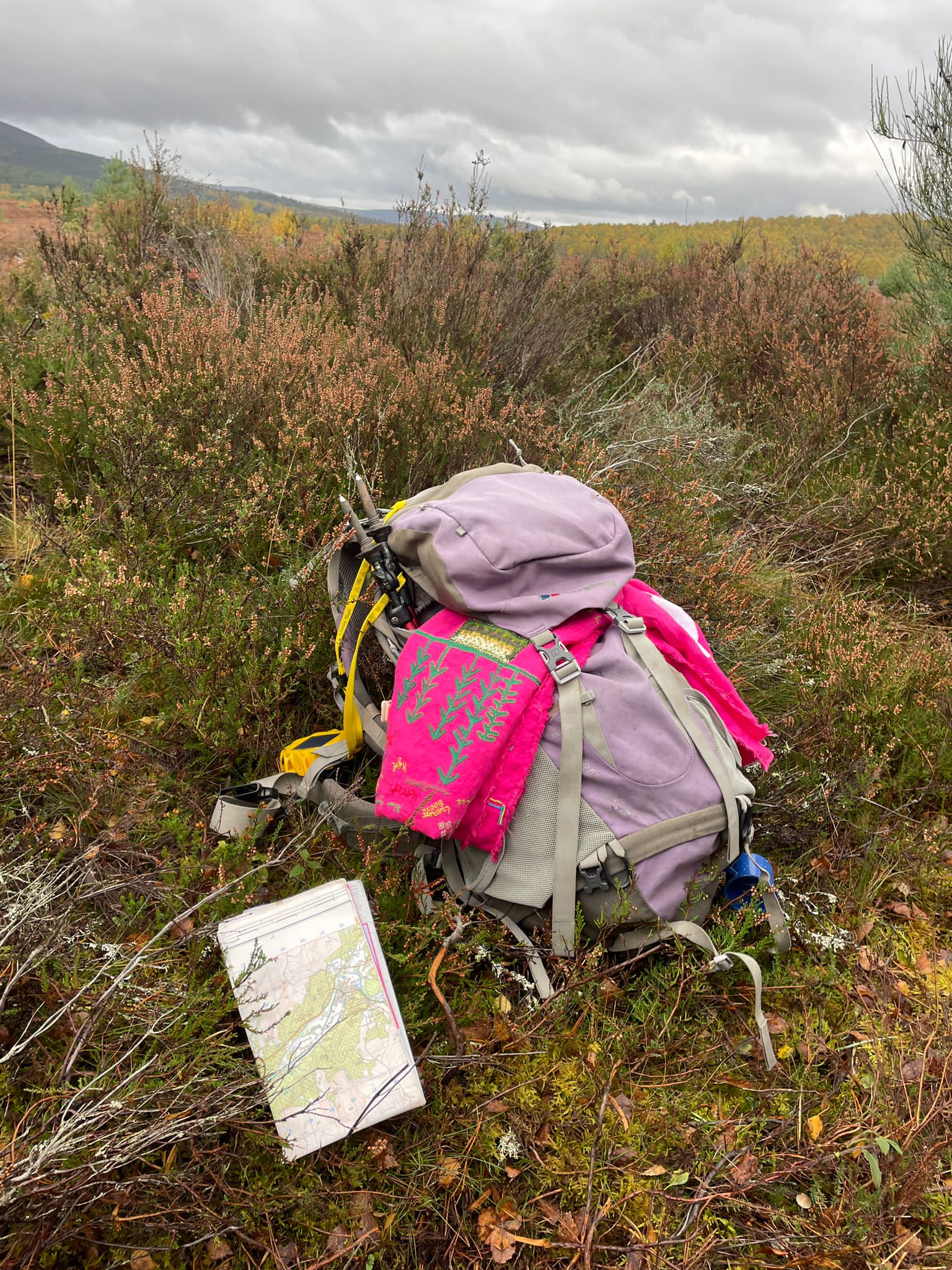 Rucksack with embroidered pink tablecloth tucked into it, with map on ground at the side surrounded by heather